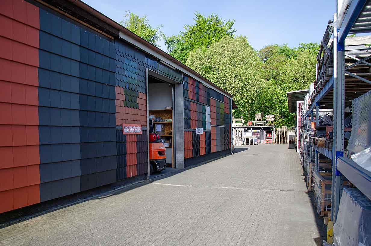 Lagerplatz mit bunten Dachziegelmustern an der Wand neben Regalen und grünen Bäumen im Hintergrund.