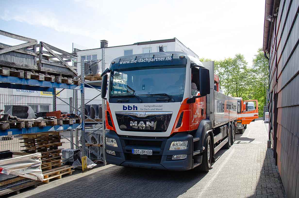 Ein MAN-LKW für Dach- und Wandbedarf der Firma bbh steht zwischen Lagerregalen und einem Gebäude.