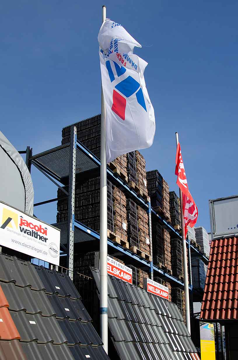 Dachziegel auf Displays und Fahnen mit Markenlogos vor Lagerregal unter blauem Himmel.