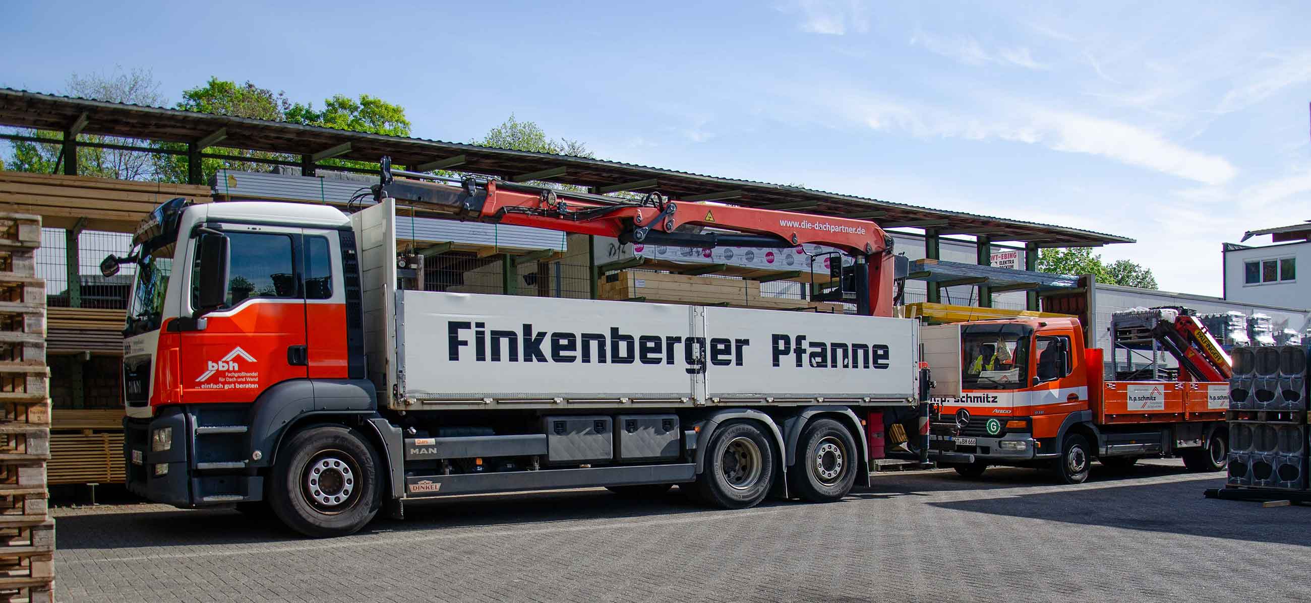 Zwei orange-weiße LKWs mit Ladekranen stehen am Lager mit Baustoffen unter einem überdachten Bereich.