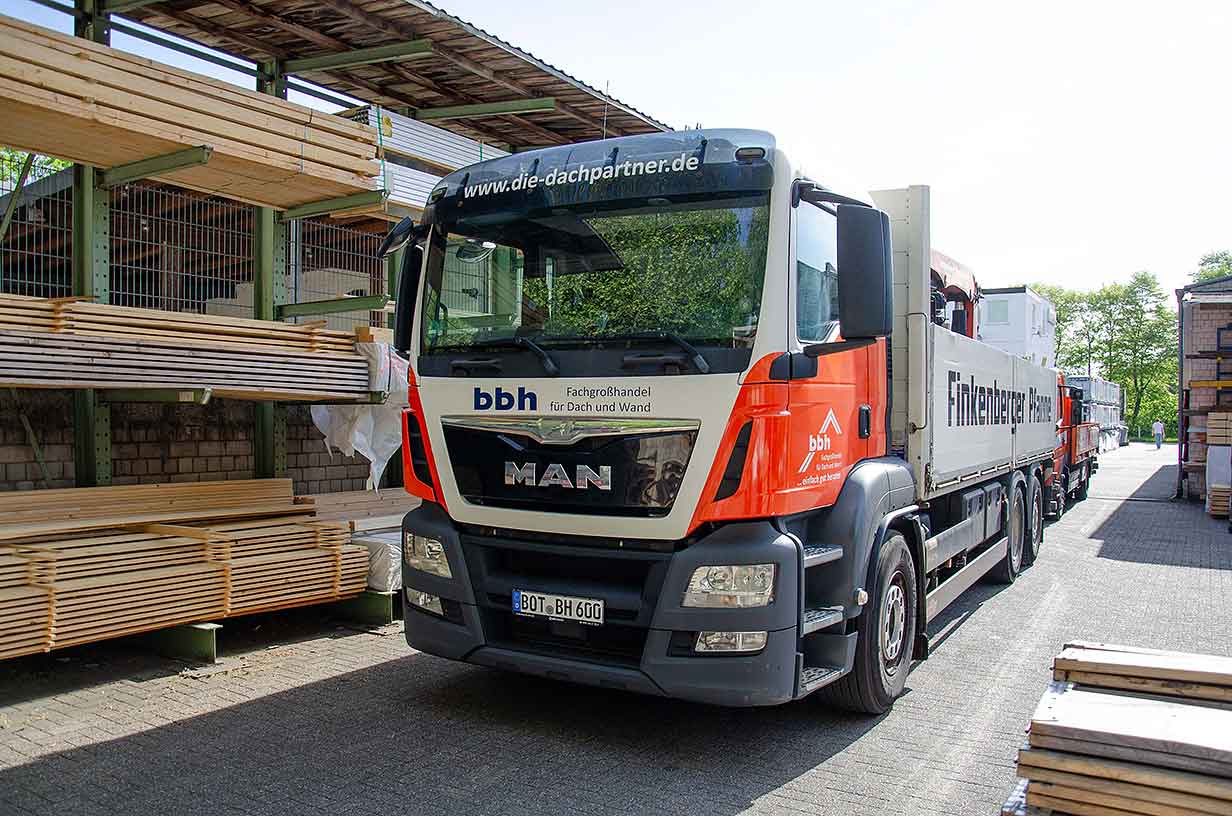 LKW der Firma bbh mit Anhänger vor einem Lager mit Holzlatten in der Sonne.