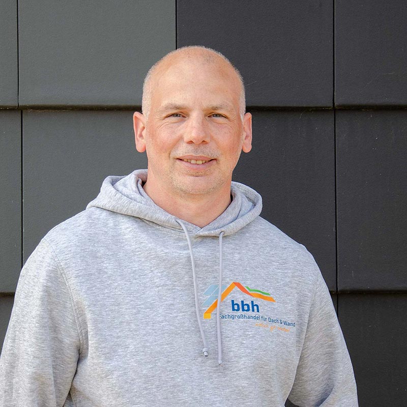 Mann in grauem Kapuzenpullover mit bbh-Logo lächelt vor schwarzer Wand.