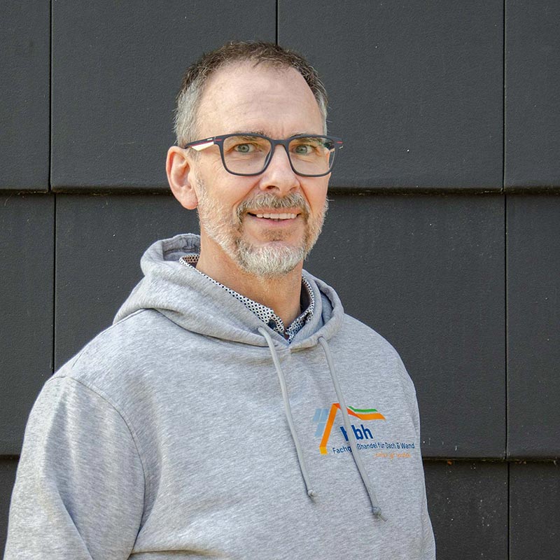 Mann mit grauem Kapuzenpullover und Brille lächelt vor schwarzer Wand.
