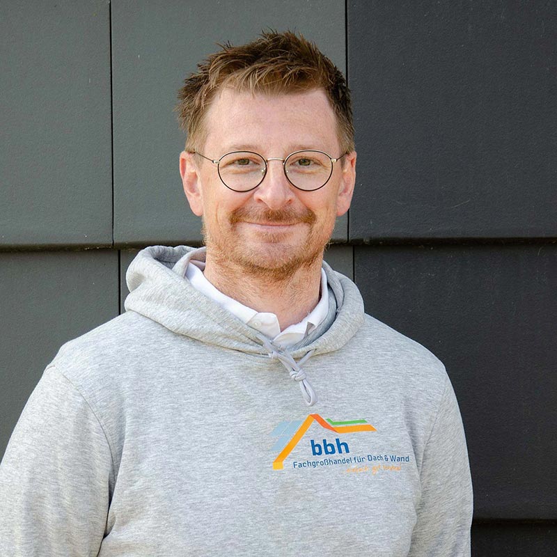 Mann mit Brille und grauem Hoodie mit bbh-Logo vor dunkler Wand