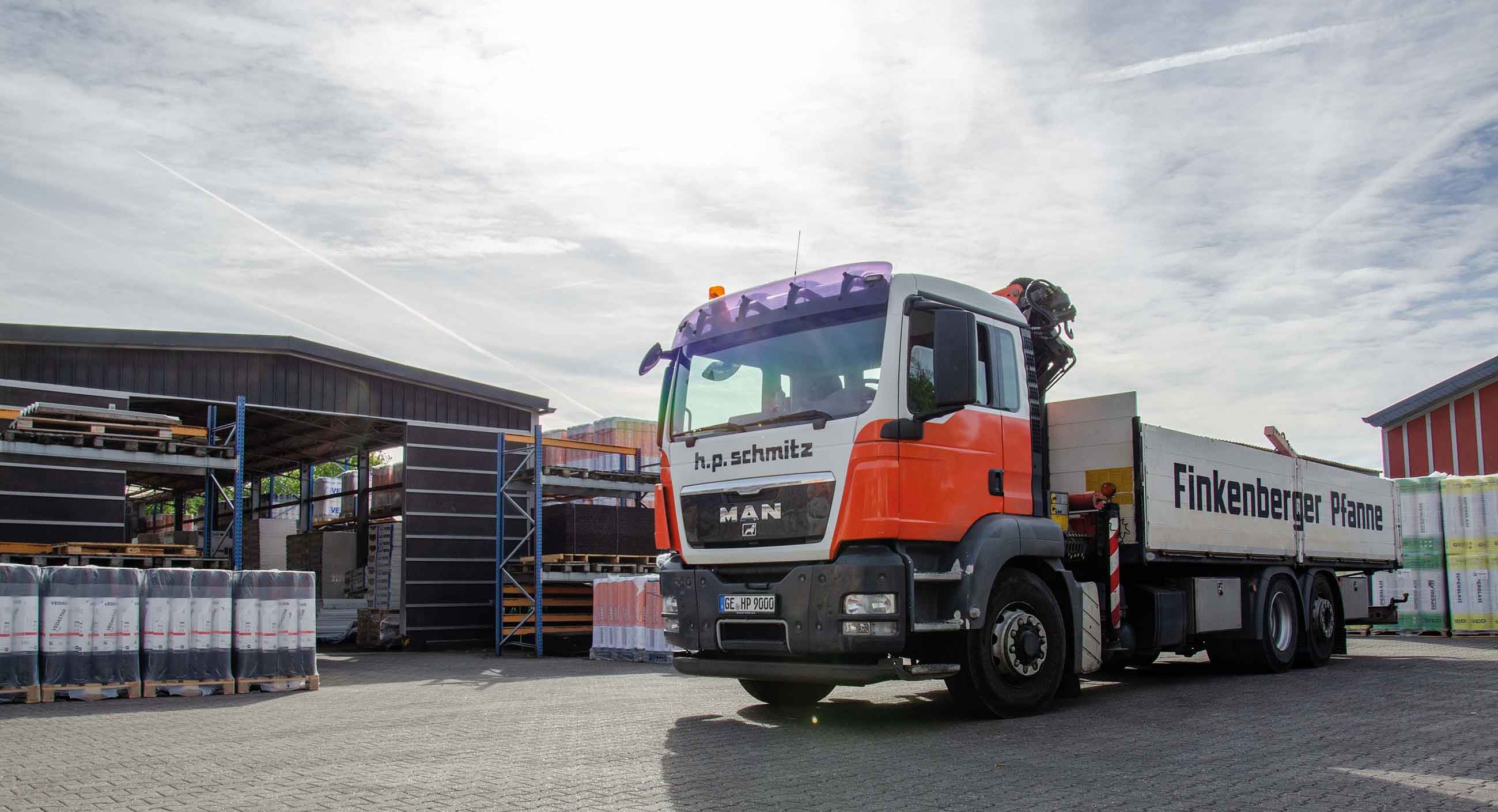 LKW mit Aufschrift h.p.schmitz und Finkenberger Pfanne neben Lager mit Baumaterialien.