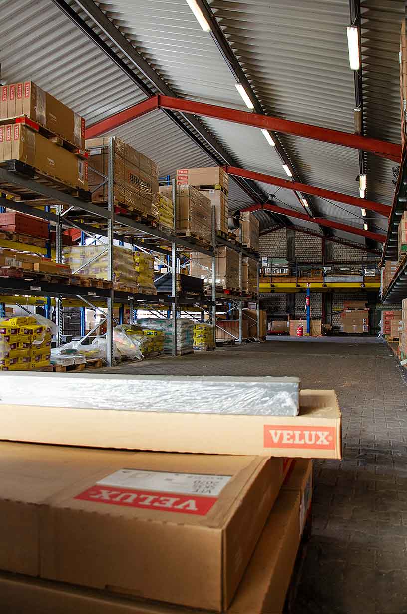 Lagerhalle mit hohen Regalen voller Kartons und verpackter Waren unter einem Dach mit roten Trägern.