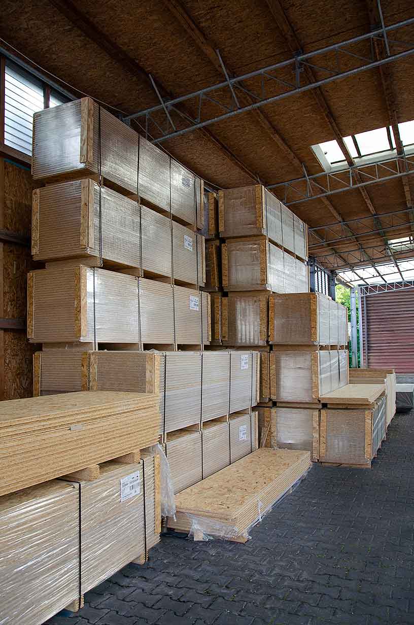Gestapelte Holzplatten in einem Lagerhaus mit sichtbarer Dachkonstruktion und Bodenbelag aus Pflastersteinen.