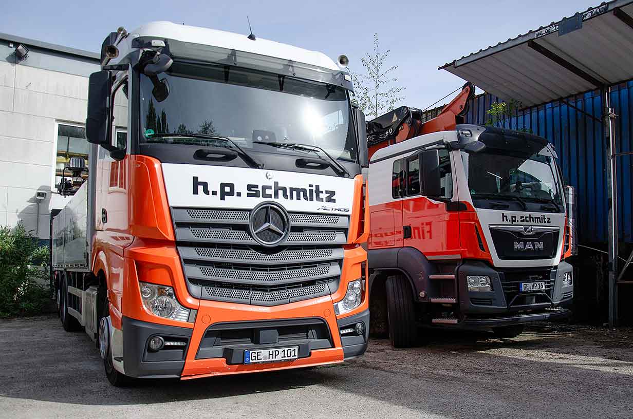 Zwei orange-weiße Lastwagen mit der Aufschrift h.p. schmitz, geparkt vor Gebäuden.