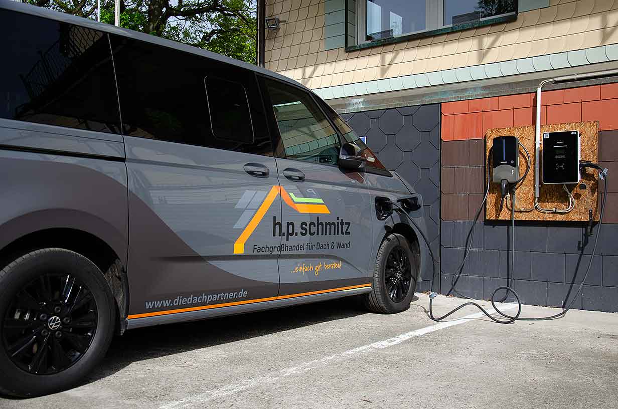 Grauer Lieferwagen von h.p.schmitz lädt an zwei Elektro-Ladestationen an einer Gebäudeaußenwand.