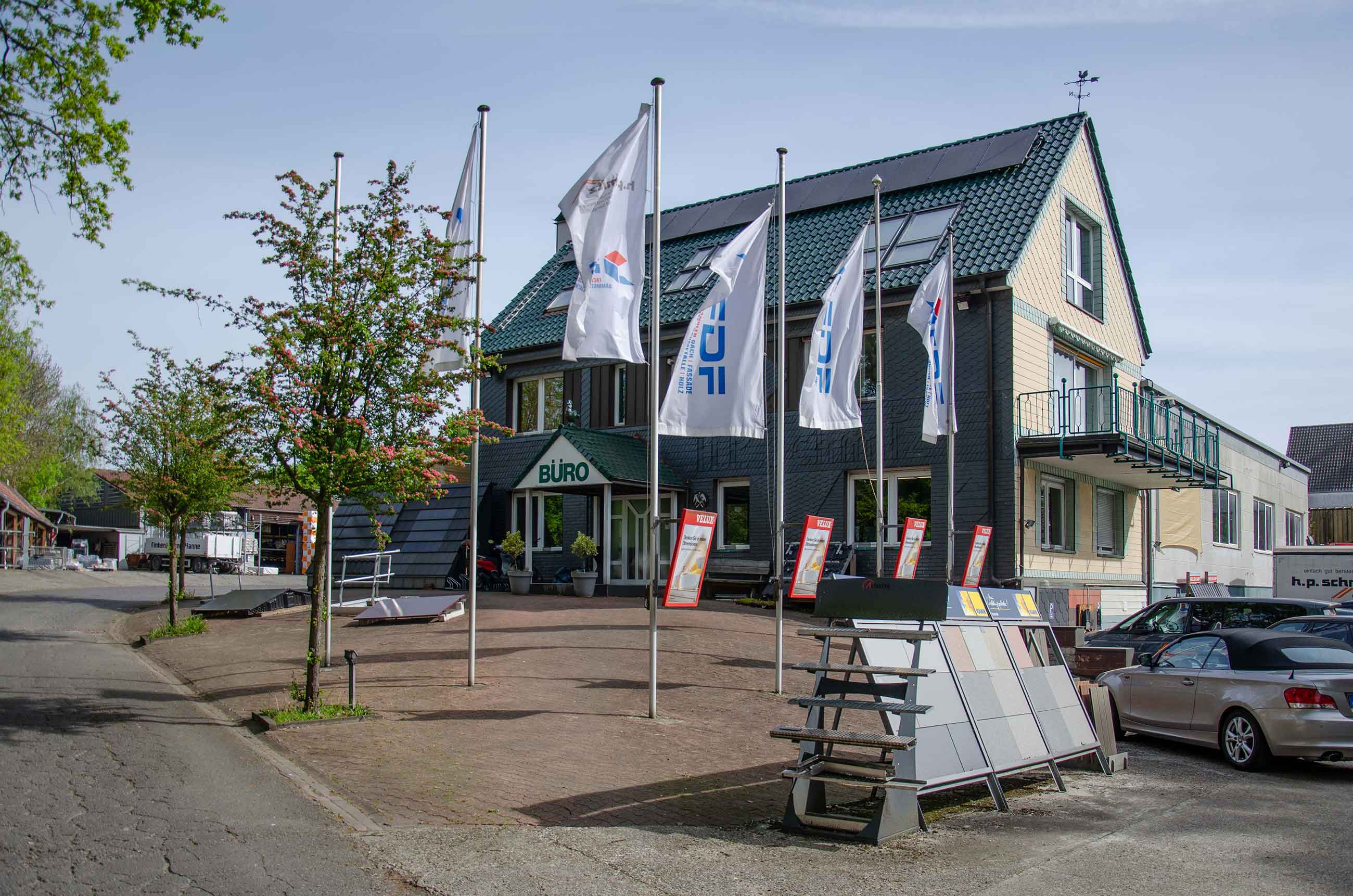 Geschäftsgebäude mit Flaggen, Pflanzen, Musterständen und geparkten Autos im Eingangsbereich.