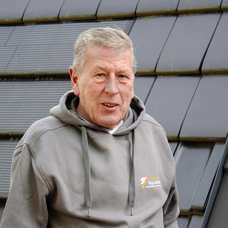 Älterer Mann mit grauem Kapuzenpullover vor einer Wand mit Solarpaneelen.