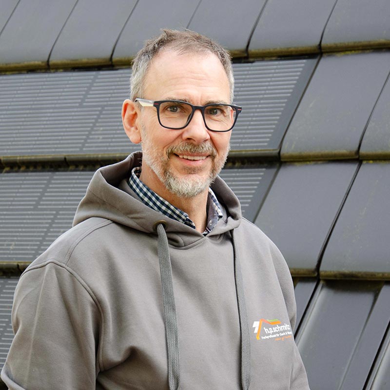 Mann mit grauem Hoodie und Brille lächelt vor einer Wand mit Solarpaneelen.