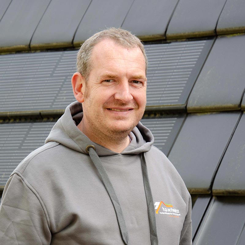 Mann in grauem Kapuzenpullover steht vor Solarzellenpaneelen und lächelt.