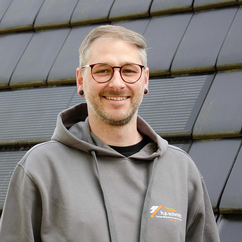Lächelnder Mann mit Brille und grauem Kapuzenpullover vor Gebäude mit dunklen Paneelen.