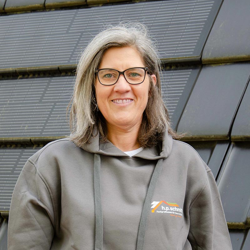Frau mit Brille und grauem Kapuzenpullover mit h.p.schmitz-Logo lächelt vor Solarpaneelen.