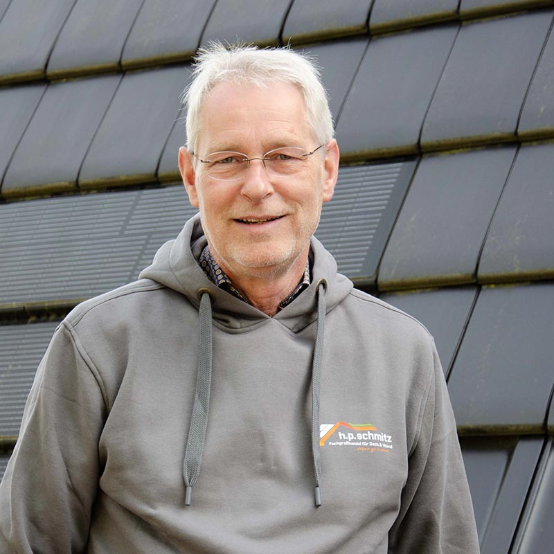 Älterer Mann mit Brille in grauem Kapuzenpullover vor dunkler Außenfassade.