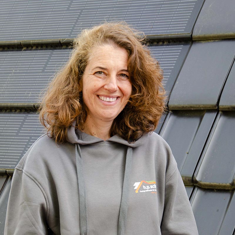 Lächelnde Frau mit lockigem Haar und grauem Kapuzenpullover vor Solarpanel-Wand.