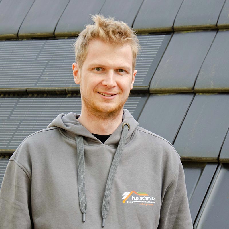 Junger Mann mit blondem Haar trägt grauen Kapuzenpullover mit h.p.schmitz Logo und lächelt.