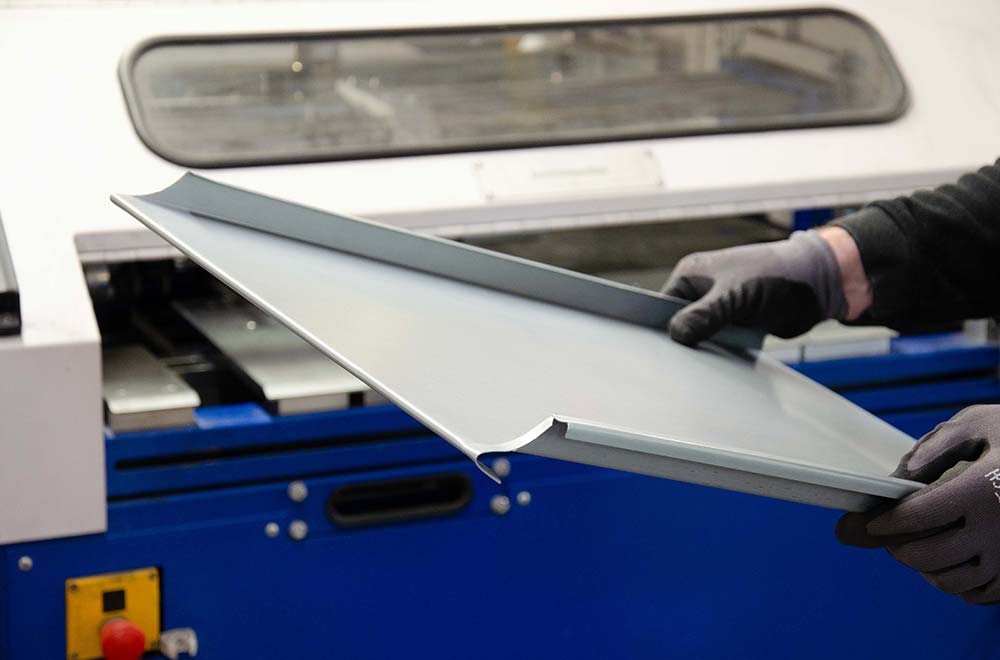 Person trägt ein geformtes Metallblech vor einer blauen Maschine in der Fabrik.