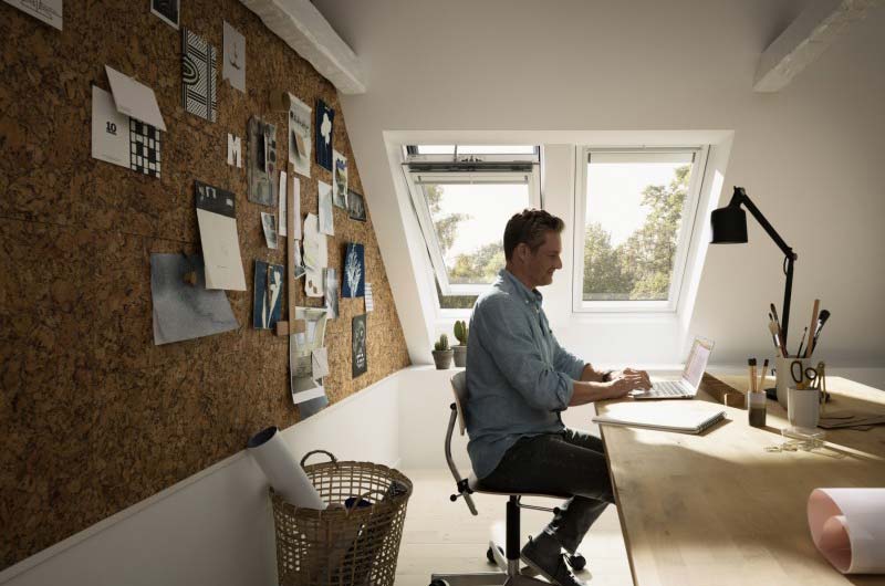 Mann sitzt an einem hellen Schreibtisch unter Dachfenstern und arbeitet an einem Laptop.